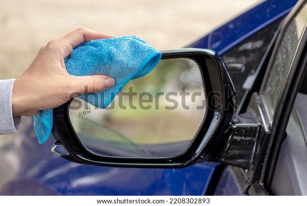 woman hand cleaning retractable rear view lateral\
car mirror with microfiber cloth dry wipe.autumn rainy day cloudy\
sky water drops on glass.sunshine sun light in mirror\
reflection.car wash\
service
