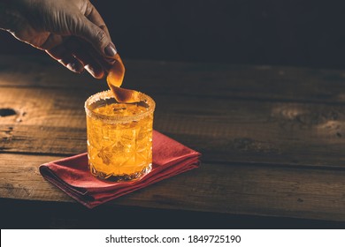 Woman Hand Decorate Orange Twist Alcoholic Cocktail Negroni On Old Wooden Board. Drink With Gin, Campari Martini Rosso And Orange, An Italian Cocktail. Nice Romantic Back Light.