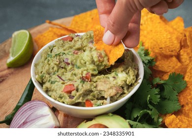 Woman hand dip nachos into tasty guacamole sauce on the wooden background. Tortilla chips next to guacamole ingredients: avocado, cilantro, onion, hot pepper and lime. Traditional Mexican food - Powered by Shutterstock