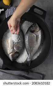 Woman Hand Dropping A Lot Of Coarse Salt Over A Pair Of Sea Breams In An Iron Cast Pan.