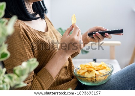 woman hand eating potato chips and holding remote tv watching series sitting on sofa 
