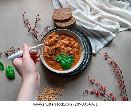 Woman hand eating vegetable lentil soup with carrot, onion, potatoes and chickpeas decorated on natural eco brown background. Hand holding spoon. Homemade cheap easy soup with fresh basil. Top view.