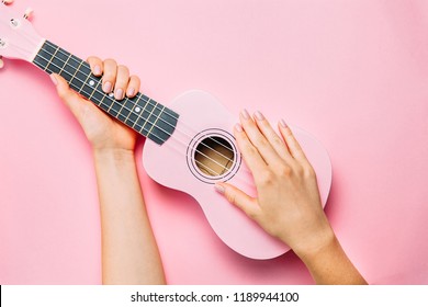 Woman hand with fashion manicure holding little pink ukulele on pink background. Fine female musician picture. Top view, flat lay.