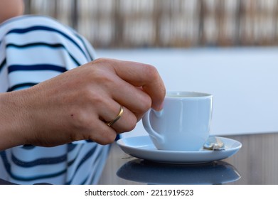 A Woman Hand With Gold Rings Holds A Cup Of Coffee From A White Saucer On A Gray Glass Table In An Outdoor Terrace