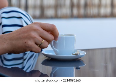 A Woman Hand With Gold Rings Holds A Cup Of Coffee From A White Saucer On A Gray Glass Table In An Outdoor Terrace