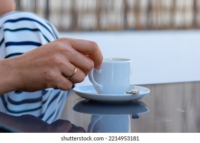 A Woman Hand With Gold Rings Holds A Cup Of Coffee From A White Saucer On A Gray Glass Table In An Outdoor Terrace