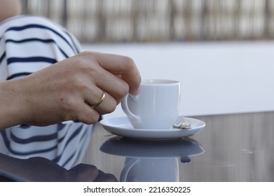 A Woman Hand With Gold Rings Holds A Cup Of Coffee From A White Saucer And A Teaspoon On A Gray Glass Table In An Outdoor Terrace