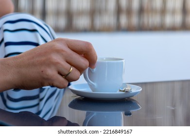 A Woman Hand With Gold Rings Holds A Cup Of Coffee From A White Saucer And A Teaspoon On A Gray Glass Table In An Outdoor Terrace