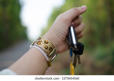 A Woman Hand Is Grabbing Keys Attached With Key Chain, Wearing Gold Bracelet Which Is Decorated With Crystals And Golden Color Design With Blurred Background Of The Road Sided With Green Forest