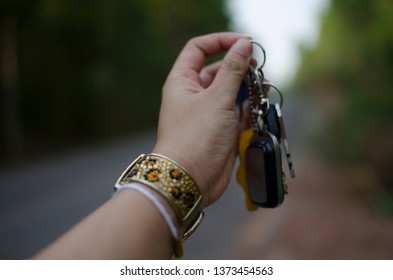 A Woman Hand Is Grabbing Keys Attached With Key Chain, Wearing Gold Bracelet Which Is Decorated With Crystals And Golden Color Design With Blurred Background Of The Road Sided With Green Forest