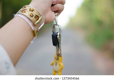 A Woman Hand Is Grabbing Keys Attached With Key Chain, Wearing Gold Bracelet Which Is Decorated With Crystals And Golden Color Design With Blurred Background Of The Road Sided With Green Forest