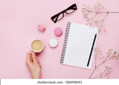 Woman Hand Hold Cup Of Coffee, Cake Macaron, Clean Notebook, Eyeglasses And Flower On Pink Table From Above. Female Working Desk. Cozy Breakfast. Flat Lay Style.