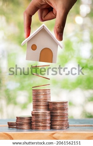 Woman hand hold a home model present to stack gold coin with growing value put on the wood in the public park, Loan or save money for buy real estate to family concept.