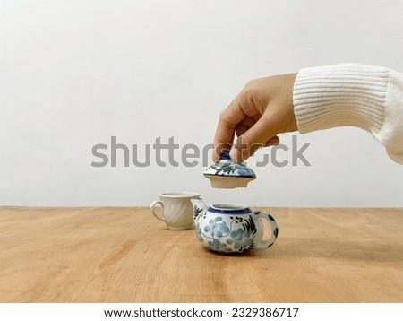 Woman hand hold mini ceramic teapot lid next to a cup standing on wooden table
