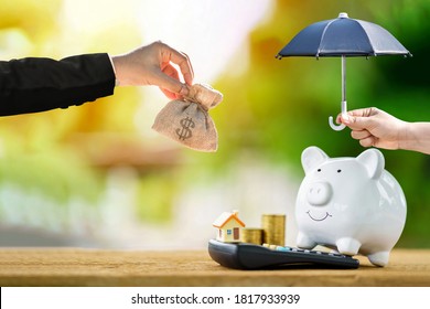 Woman hand hold open black umbrella protect the piggy bank and home and put on calculator and hold money bag in the public park, Loan or saving money for real estate and property protection concept.