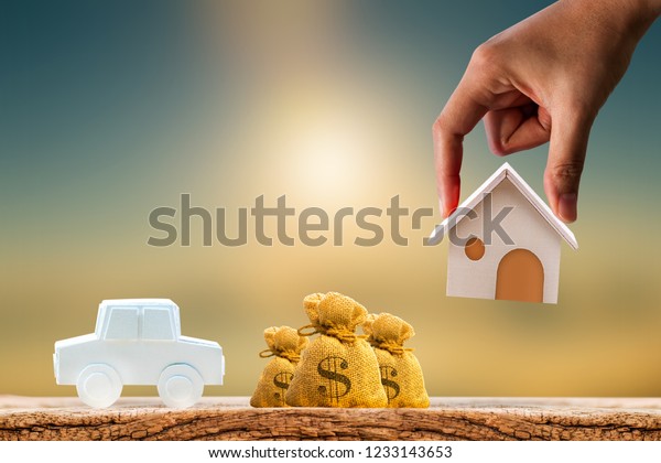 Woman hand hold\
for select a home model and money bag and car on the wood on\
sunlight, Loan for asset or saving money for buy a new real estate\
to family in the future\
concept.