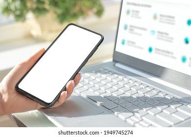 Woman Hand Holding The Black Cell Phone Smartphone With Blank White Screen And Modern Frame Less Design On Black Background. Mockup Phone. Hand Holding Mobile Phone. Mockup Cell Phone On Laptop