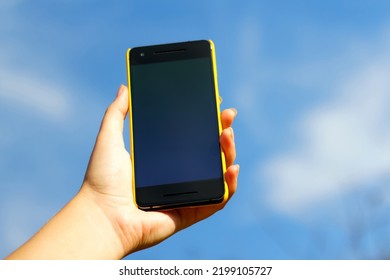 Woman Hand Holding Black Smartphone In Yellow Plastic Case  On Blue Sky Nature Background, Sunny Day. Black Screen. Mockup, Mock-up, Mock Up. Female Hand. Smart Phone.