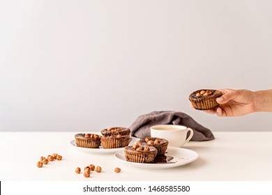 Woman Hand Holding A Chocolate Cupcake With Walnuts. Chocolate Cupcakes And A Cup Of Coffee On White Table Front View