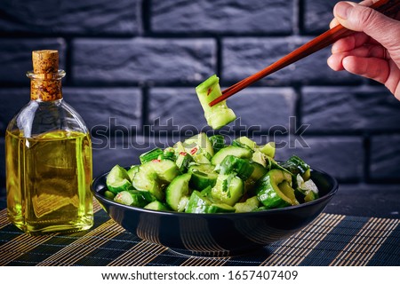 woman hand is holding chopsticks with a cucumber chunk of Chinese Smashed Cucumber salad, pai huang gua in a bowl on a concrete table with a brick wall at the background, close-up, horizontal view