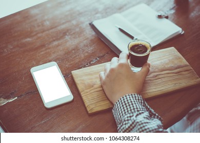Woman hand holding cup of coffee with smartphone, pen, note page on wooden table. Top view.