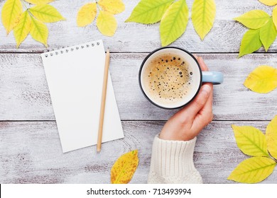 Woman Hand Holding Cup Of Coffee, Open Notebook And Autumn Leaves On Vintage Wooden Table Top View. Cozy Breakfast. Fall Bucket List. Flat Lay