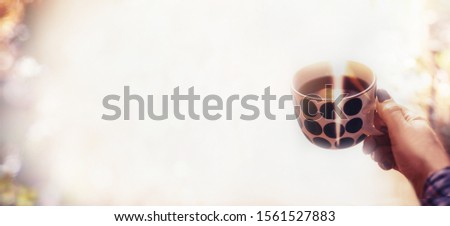 Woman hand holding cup of hot chocolate at blurred background with sunlight bokeh. Wide composition. Copy space