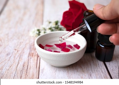 Woman of hand holding dropper and dripping oil into white bowl.Brown bottles,red rose and white cutter flowers on white wooden table.Aromatherapy and essential organic concept. - Shutterstock ID 1548194984
