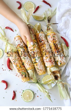 a woman hand holding Elote, grilled Mexican street Corn, Summer, BBQ , fast, healthy food concept, top view, flat lay, white background, decorated with red hot chili peppers lime and cilantro leaves