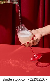 Woman hand holding glass and pouring wine of bottle. Woman tasting white wine on red background. Minimal trendy style composition with person and champagne. Pouring wine and female hand with glass