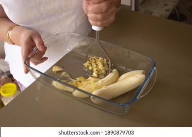 woman hand holding a glass pyrex full of banana slices while smashes the fruit inside using a kitchen tool, closeup.