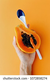 Woman Hand Holding Half Hawaii Papaya And Spoon On An Orange Background, Food Concept Top View