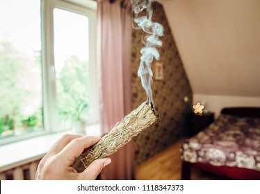 Woman Hand Holding Herb Bundle Of Dried Sage Smudge Stick Smoking. It Is Believed To Cleanse Negative Energy And Purify Living Spaces At Home In Rooms.