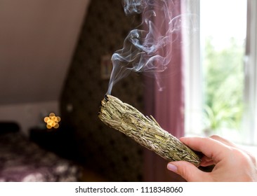 Woman Hand Holding Herb Bundle Of Dried Sage Smudge Stick Smoking. It Is Believed To Cleanse Negative Energy And Purify Living Spaces At Home In Rooms.