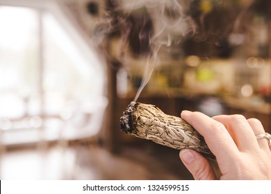Woman hand holding herb bundle of dried sage smudge stick smoking. It is believed to cleanse negative energy and purify living spaces at home in rooms.