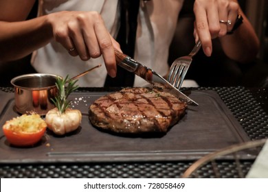Woman hand holding knife and fork cutting grilled beef steak on stoned plate eat with steak side dish and sauce for dinner meal in restaurant - selective focus
