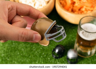 Woman Hand Holding A Little American Football Helmet. Snacks, Little Helmets And Beer On Green Grass Background. American Football Decoration