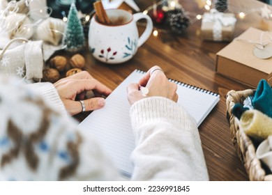 Woman hand holding pen on notepad at home on winter holidays xmas. Goals plans make to do and wish list for new year christmas concept, girl writing in notebook. Christmas decoration, gift boxes. 
