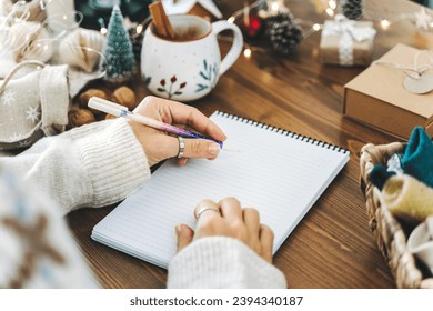 Woman hand holding pen on notepad at home on winter holidays xmas. Goals plans make to do and wish list for new year christmas concept, girl writing in notebook. Christmas decoration, gift boxes. 