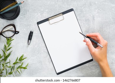 Woman hand holding pen and write a message in blank paper clipboard. Feminine home office desk with stationery, glasses, branch with green leaves on concrete stone background. Business concept.
