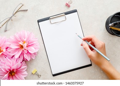 Woman Hand Holding Pencil And Writing A Text Into Blank Paper Clipboard Mockup. Flat Lay Home Office Desk. Top View Female Workspace With Paper Clipboard Mockup, Pink Dahlia Flowers, Woman Accessories