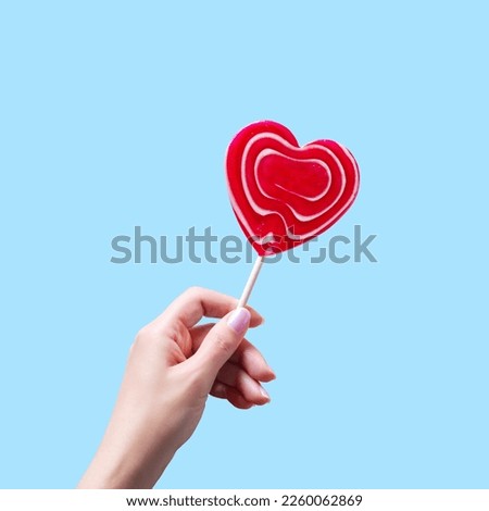 Woman hand holding red heart lollipop on blue background, card for Valentines Day celebration.