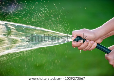 Woman hand holding rubber water hose and using finger close end of rubber water hose to make  water spray with sunlight and green grass field in background. World Water Day conpect.