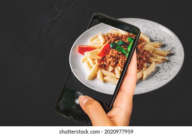 woman hand holding and showing smart phone takes a photo Pasta penne bolognese in white plate on wooden table background. food photos
