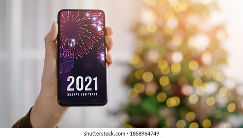 Woman Hand Holding Smartphone With Happy New Year 2021 Word On Screen With Light Bokeh Background. Christmas Mockup With Space For Text.