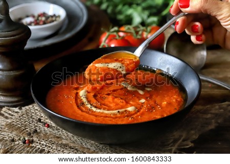 Woman hand holding a spoon full of tomato soup on wooden rustic background with herbs and cherry tomatoes in the back. Italian dinner or lunch tomato soup, horizontal photo