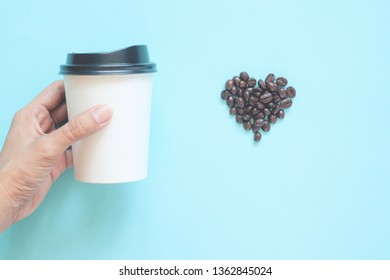 Woman Hand Holding Takeaway Coffee Cup With Heart Shape Coffee Beans On Pastel Colour Background. Coffee Poster Advertisement