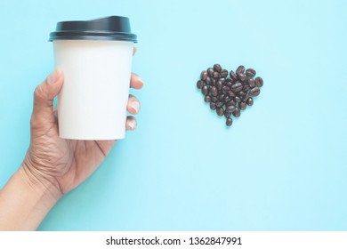 Woman Hand Holding Takeaway Coffee Cup With Heart Shape Coffee Beans On Pastel Colour Background