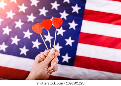 Woman Hand Holding Three Red Hearts Shape On Stick In Front Of American Flag. Visual Concept Of Preparation For Independence Day. Fourth Of July Patriotic Consept.