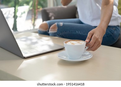 Woman Hand Holding White Coffee Cup With Blurry Labtop, Desk And Sofa In Cafe Background. Girl Working Online Business Concept At Home Or Cafe.